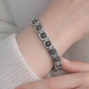 May include: Silver-toned bracelet featuring olive-green stones and small clear accents. The bracelet is worn on a wrist, showcasing its design and detailing. The jewelry is a stylish accessory.