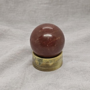 May include: A polished, reddish-brown sphere, possibly a gemstone, sits atop a small, weathered, circular stand. The sphere has subtle surface variations and a glossy finish. The stand is a muted gold color.