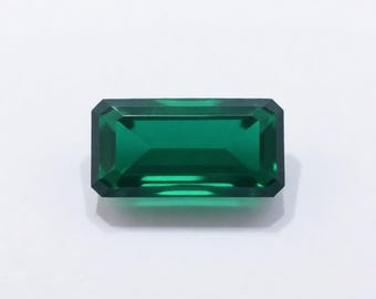 Puede incluir: Una gema rectangular con talla esmeralda de color verde intenso. La gema tiene una superficie facetada que refleja la luz, creando un brillo brillante. Los bordes están bien definidos y la forma general es simétrica. La gema está sobre un fondo blanco.