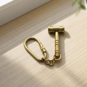 May include: A brass-colored miniature hammer keychain. The hammer head is cylindrical, and the handle is ribbed. The keychain includes a split ring and a clasp. The item is on a light-colored wooden surface.
