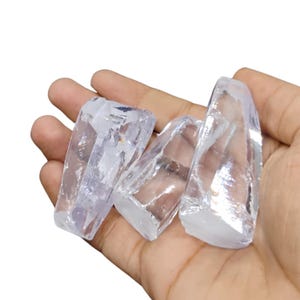 May include: Four clear quartz crystal pieces with a translucent appearance are held in a hand. The crystals have a rough, natural texture and varying shapes. The light refracts through the quartz, creating highlights and shadows.