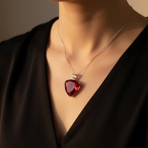 May include: A silver necklace with a large, triangular, deep red gemstone pendant. The pendant is set in a silver bezel and hangs from a delicate silver chain. The necklace is worn against a black garment.