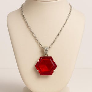 May include: A silver necklace with a hexagonal, red pendant. The pendant is a faceted, gem-like shape. The necklace is displayed on a cream-colored bust.