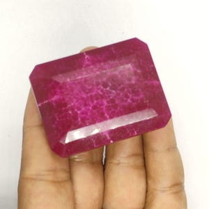 Peut inclure: Une grosse pierre précieuse rectangulaire et facettée, de couleur magenta foncé. La gemme présente un intérieur texturé et une surface polie et réfléchissante. Les bords sont biseautés, et la pierre est tenue dans une main sur fond blanc.