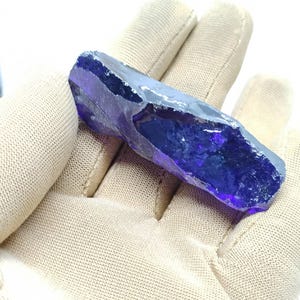 May include: A striking, raw crystal in a deep blue hue, held in a beige fabric glove. The crystal's surface is faceted, with a rough texture along the edges. The elongated shape and intense colour make it a visually appealing specimen.