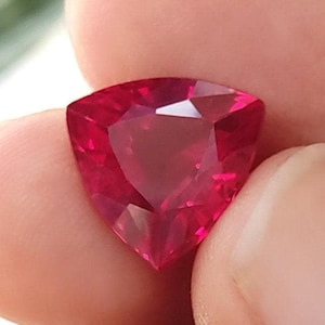 May include: A triangular, faceted, deep red gemstone. The gem has a brilliant cut, reflecting light across its many facets. The stone is held between fingers, showcasing its vibrant colour and clarity. The gem appears to be a ruby.