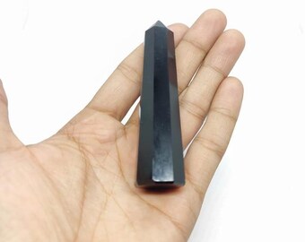 May include: A polished black obsidian obelisk, a gemstone with a faceted, elongated shape. The dark, opaque stone is held in a hand against a white background. The obelisk has a flat base and a pointed top.
