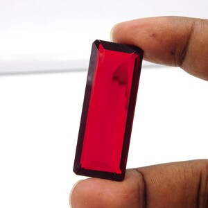 May include: A rectangular, faceted, deep red gemstone with a dark border. The gem is held between two fingers against a white background. The gem's facets catch the light, creating a brilliant display of color.