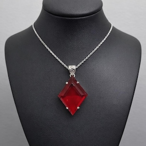 Op de afbeelding: Zilveren ketting met een grote, facetgeslepen, rode ruitvormige hanger. De hanger heeft een diepe robijnrode kleur en is gezet in een zilveren metalen frame. De ketting wordt getoond op een zwarte mannequin.