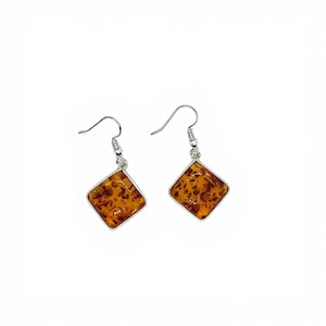 May include: A pair of dangle earrings with a square, amber-colored center. The earrings have a silver-colored metal frame and hook. The amber-colored center has a speckled pattern of brown and orange.