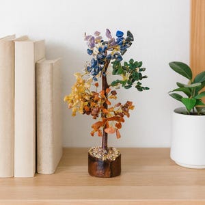 Pode incluir: Uma escultura de árvore de pedras preciosas com um tronco castanho e uma base de madeira. As folhas da árvore são feitas de pedras preciosas de várias cores, incluindo roxo, azul, verde, amarelo e laranja. A escultura é exibida numa prateleira de madeira com livros e uma planta em vaso.