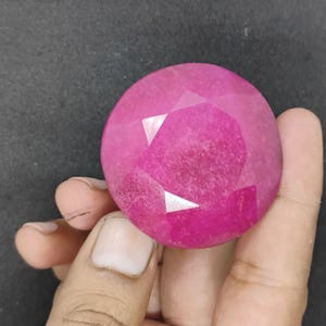 May include: A large, round ruby gemstone with multiple facets. The gem is a vibrant pinkish-red colour, with the facets creating a sparkling effect. The stone is held in a hand against a dark background.