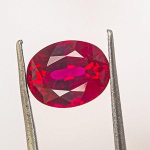 May include: A faceted, oval-shaped ruby gemstone held by metal tweezers. The ruby displays a deep red color with a brighter, more saturated center. The gemstone's cut reveals multiple facets, reflecting light.