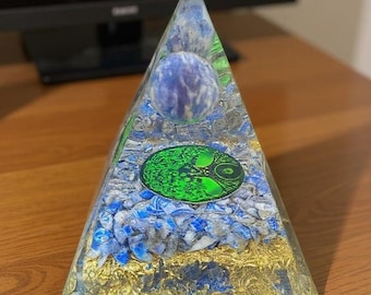 May include: A clear resin pyramid with a blue and white orb at the top. The pyramid contains blue and gold flecks, and a green tree of life symbol. The pyramid sits on a wooden surface.