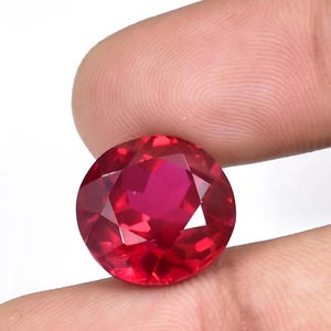 May include: A faceted, round-cut ruby gemstone. The gem is a deep, vibrant red color with multiple facets that reflect light. The ruby is held between two fingers against a white background.