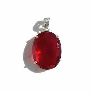 May include: A red oval gemstone pendant with a silver-coloured bail. The gemstone is a deep, vibrant red colour and is set in a silver-coloured metal setting. The pendant is displayed against a white background.