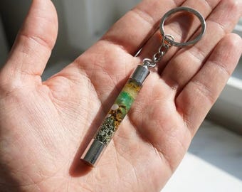May include: A handmade keychain featuring a cylindrical pendant filled with natural elements. The pendant has silver-colored metal caps and a silver key ring. The interior of the pendant displays green, brown, and white organic materials.