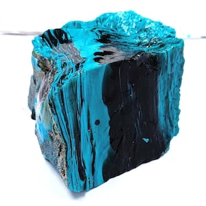 Puede incluir: Un bloque de piedra con una superficie rugosa y texturizada, pintado con una llamativa combinación de turquesa y negro. Los colores están en capas y arremolinados, creando un efecto veteado. La piedra es un artículo decorativo único.