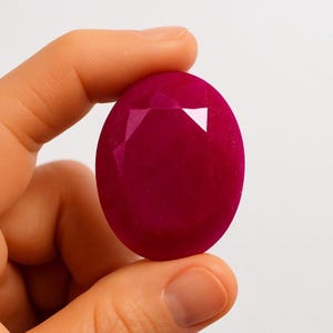 May include: A large, oval-cut ruby gemstone. The gem is a deep, saturated red color with facets that catch the light. The stone is held between fingers, showcasing its size and brilliance. The background is a plain white.