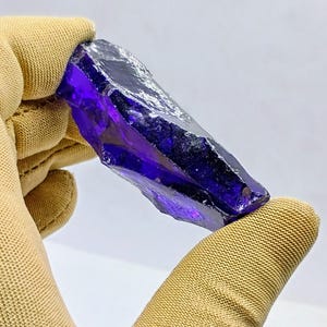 Pode incluir: Um cristal bruto, sem corte, de cor roxa escura, segurado por uma luva bege. O cristal tem uma forma irregular e irregular com uma qualidade translúcida. A cor varia de escura, quase preta, a um violeta vibrante.
