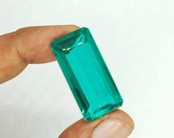 Puede incluir: Una gema rectangular de color verde esmeralda con una superficie facetada. La gema se sostiene entre dos dedos sobre un fondo blanco, mostrando su claridad y color. El corte de la piedra permite que la luz se refleje, creando un efecto brillante.