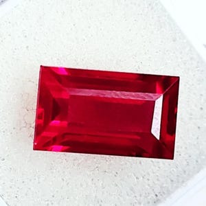Può includere: Una gemma rettangolare, sfaccettata, di colore rosso intenso. La gemma ha una superficie brillante e lucida con bordi e angoli vivi. Il colore è un rosso ricco e saturo, con un alto grado di chiarezza. La gemma è posta su uno sfondo bianco.