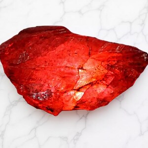 May include: A vibrant, irregularly shaped, red gemstone with a textured surface. The gemstone is set against a white marble background, highlighting its rich color and natural form. The stone's surface reflects light, creating a shimmering effect.