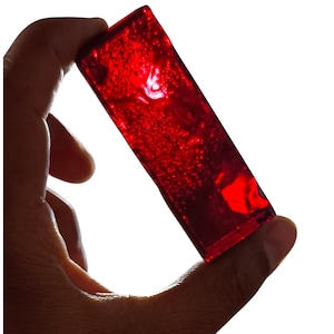May include: A rectangular, translucent, red object is held against a white background. The object appears to be made of a shiny material, with a textured surface. The light shines through the object, creating a glowing effect.