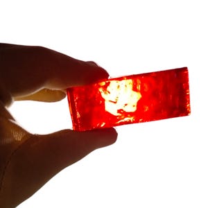 Puede incluir: Un objeto rectangular de vidrio rojo translúcido se sostiene sobre un fondo blanco. El objeto está iluminado, creando un efecto brillante y luminoso. Los bordes del vidrio son ligeramente irregulares.