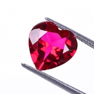 Könnte beinhalten: Ein herzförmiger, facettierter, rubinroter Edelstein wird von einer silbernen Pinzette gehalten. Die polierten Oberflächen des Edelsteins reflektieren das Licht und erzeugen ein brillantes Farb- und Funkeln. Der Edelstein hat einen leuchtenden Rotton.