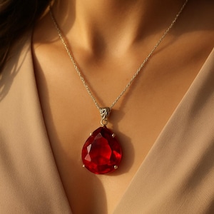 Puede incluir: Un collar de plata con un colgante grande en forma de lágrima. El colgante es una gema roja facetada y vibrante, engastada en un bisel de plata. El collar se muestra sobre un fondo neutro.