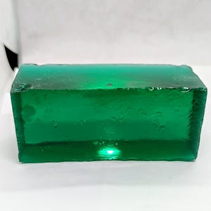 Könnte beinhalten: Ein rechteckiger Block aus smaragdgrünem Glas mit unregelmäßigen Kanten. Das durchscheinende Material lässt Licht durch, wodurch ein Leuchteffekt entsteht. Der Block steht vor einem weißen Hintergrund.