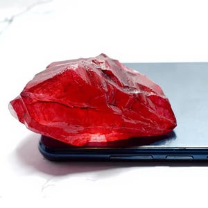 Peut inclure: Une grosse gemme rouge foncé, de forme irrégulière, repose sur un smartphone bleu foncé. La gemme a une surface rugueuse et texturée et semble translucide. Le fond est une surface blanche brillante.