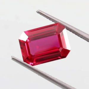 May include: A faceted, rectangular ruby gemstone held by silver tweezers. The ruby has a deep red color with sharp, clean edges and a polished surface, reflecting light. The gemstone is set against a plain white background.