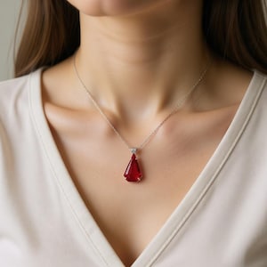 May include: A silver necklace with a triangular, faceted, deep red pendant. The pendant is suspended from a delicate chain and rests against a light-colored top. The gemstone has a brilliant cut, catching the light.
