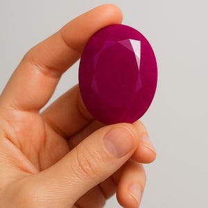 Op de afbeelding: Een gefacetteerde, ovaalvormige edelsteen in een diepe magenta tint. De edelsteen heeft een gepolijst oppervlak met meerdere facetten die het licht vangen, waardoor een sprankelend effect ontstaat. De edelsteen wordt in een hand gehouden.
