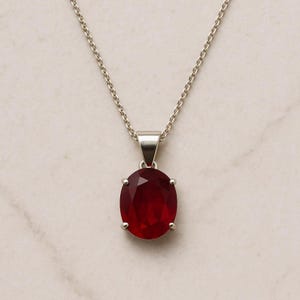 May include: A silver necklace with a large, oval-shaped, deep red gemstone pendant. The pendant is secured by four silver prongs and suspended from a fine silver chain. The gemstone's faceted surface catches the light.