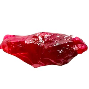 Op de afbeelding: Een ruwe, ongeslepen robijn edelsteen met een dieprode kleur. De ruwe steen heeft een onregelmatige vorm en een enigszins doorschijnend uiterlijk, dat licht over het oppervlak reflecteert. De robijn staat tegen een effen witte achtergrond.