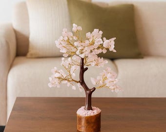 Könnte beinhalten: Ein dekorativer Rosenquarz-Kristallbaum mit einem braunen Stamm und goldfarbenen Drahtzweigen. Der Baum steht auf einem Holzsockel und hat hellrosa Kristallblätter. Der Baum steht auf einem dunkelbraunen Holztisch.