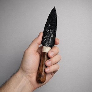 May include: A black obsidian knife with a wooden handle. The handle is wrapped with a natural-colored twine. The blade is a dark, glossy black, and the handle is a warm brown color. The knife is held in a hand against a gray background.