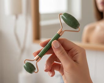 Puede incluir: Un rodillo facial de jade verde con detalles en oro rosa. El rodillo tiene dos piedras de jade redondeadas conectadas por un marco de oro rosa y un mango verde. El mango también está acentuado con oro rosa. El producto se sostiene en una mano.