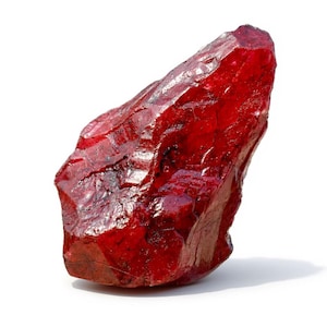 Könnte beinhalten: Ein grob geschliffener, tiefroter Edelstein mit glänzender, reflektierender Oberfläche. Das rubinrote Mineral hat eine gezackte, unregelmäßige Form und ist vor einem weißen Hintergrund platziert. Die Facetten des Steins fangen das Licht ein und betonen seine satte Farbe und Textur.