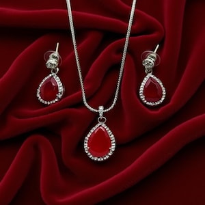 Pode incluir: Um conjunto de joias prateadas com um colar e brincos a condizer. Cada peça apresenta uma pedra vermelha escura em forma de gota, rodeada por pequenos cristais transparentes. O conjunto é exibido sobre um fundo de veludo vermelho intenso.