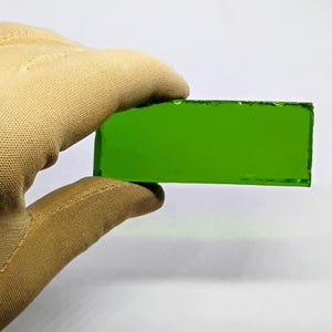 May include: A rectangular piece of bright green glass is held by a gloved hand against a white background. The glass has a smooth, polished surface and appears to be translucent. The edges of the glass are slightly rough.