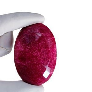 Puede incluir: Una gema facetada, de forma ovalada, en un tono rojo intenso y rico. La gema tiene una superficie pulida con vetas internas y algunas facetas que captan la luz. La gema es sostenida por una mano enguantada sobre un fondo blanco.