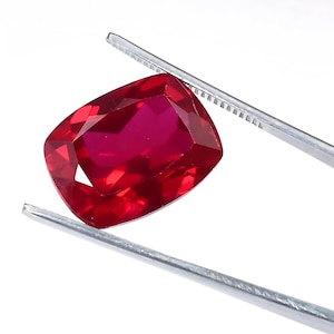 Può includere: Un rubino taglio a cuscino, di colore rosso intenso, tenuto da pinzette argentate su sfondo bianco. La gemma presenta molteplici sfaccettature che riflettono la luce. Le pinzette sono posizionate diagonalmente, evidenziando la forma e il colore del rubino.