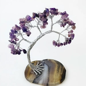 Pode incluir: Uma escultura decorativa de árvore com tronco e galhos de arame prateado, adornada com folhas de pedras preciosas de ametista roxa. A árvore é montada em uma base de ágata polida, em forma oval, com faixas marrons, brancas e cinzas.
