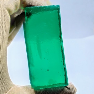 Könnte beinhalten: Ein rechteckiges, durchscheinendes, smaragdgrünes Glasstück. Die Kanten haben eine raue, unebene Textur. Das Objekt wird von einer behandschuhten Hand vor weißem Hintergrund gehalten.
