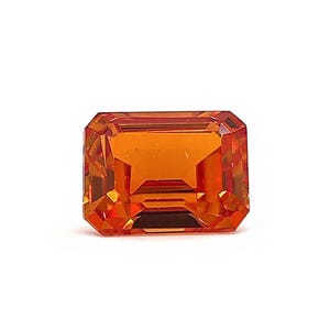 Puede incluir: Una gema naranja tallada en forma rectangular. La gema tiene múltiples facetas y está sobre un fondo blanco. Las facetas crean un juego de luces y sombras, realzando su brillo.