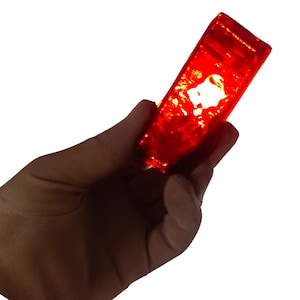 May include: A rectangular, translucent, red object is held in a hand against a white background. The object appears to be made of glass or a similar material, with light shining through it, creating a glowing effect.